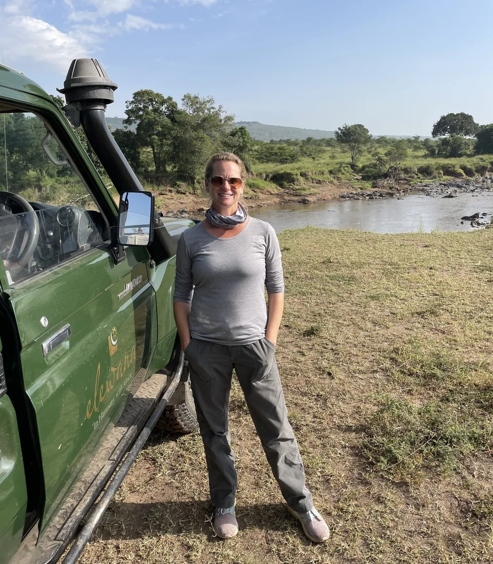 A traveler beside a green safari vehicle on a riverbank.